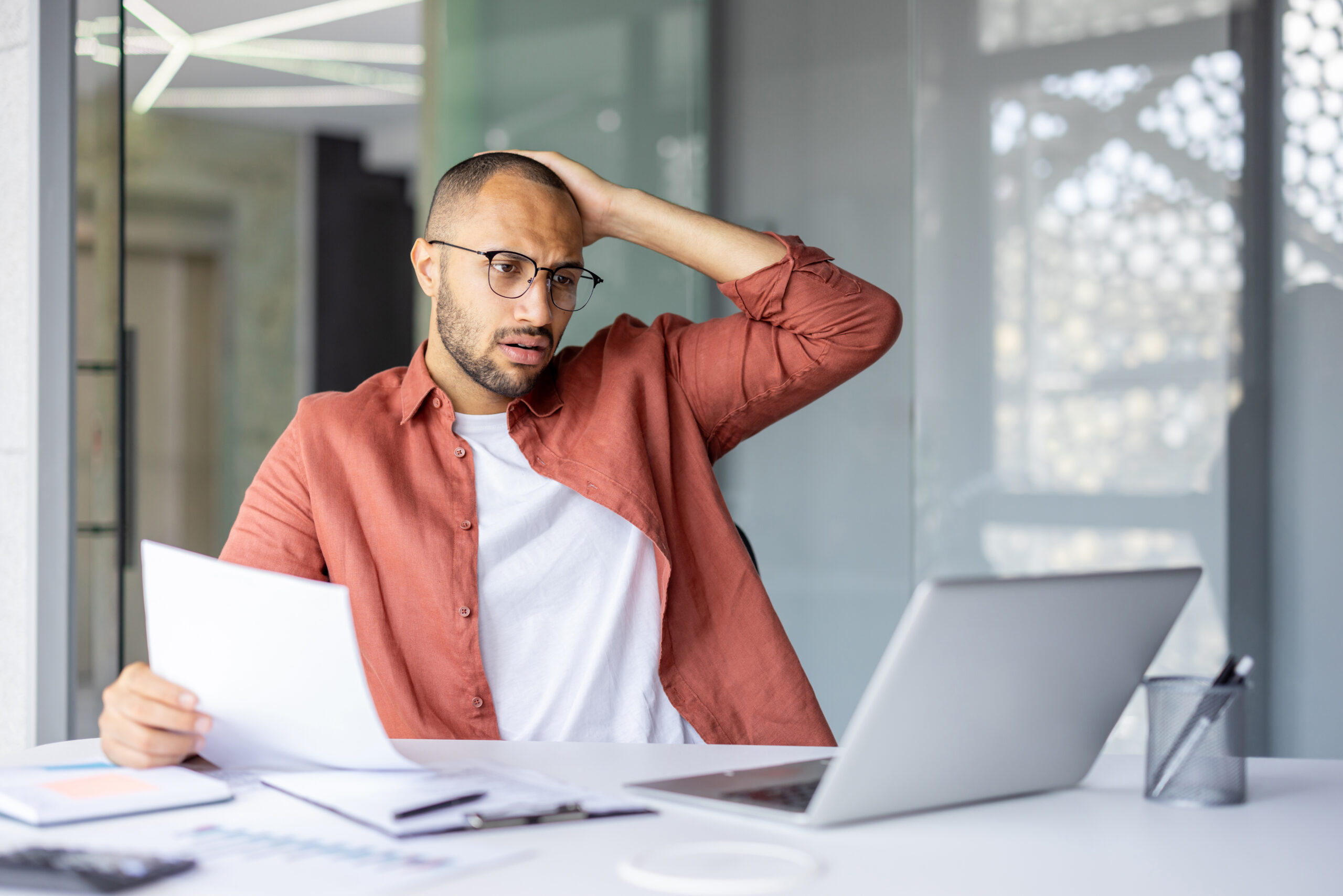 Stressed man at desk with laptop and paperwork, possibly needing umbrella coverage.