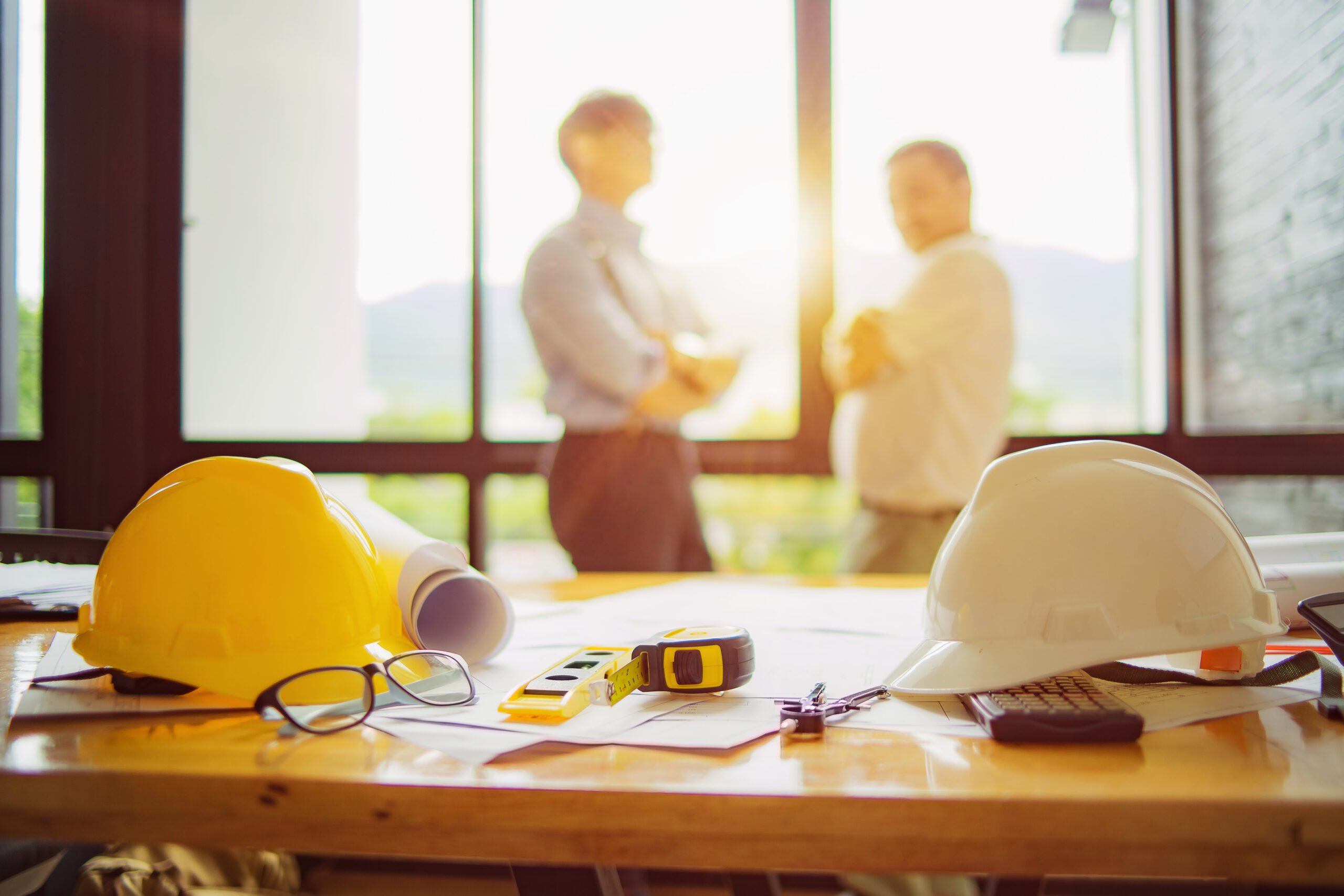 Construction plans, hard hats, and tools on a table, with two developers in the background.